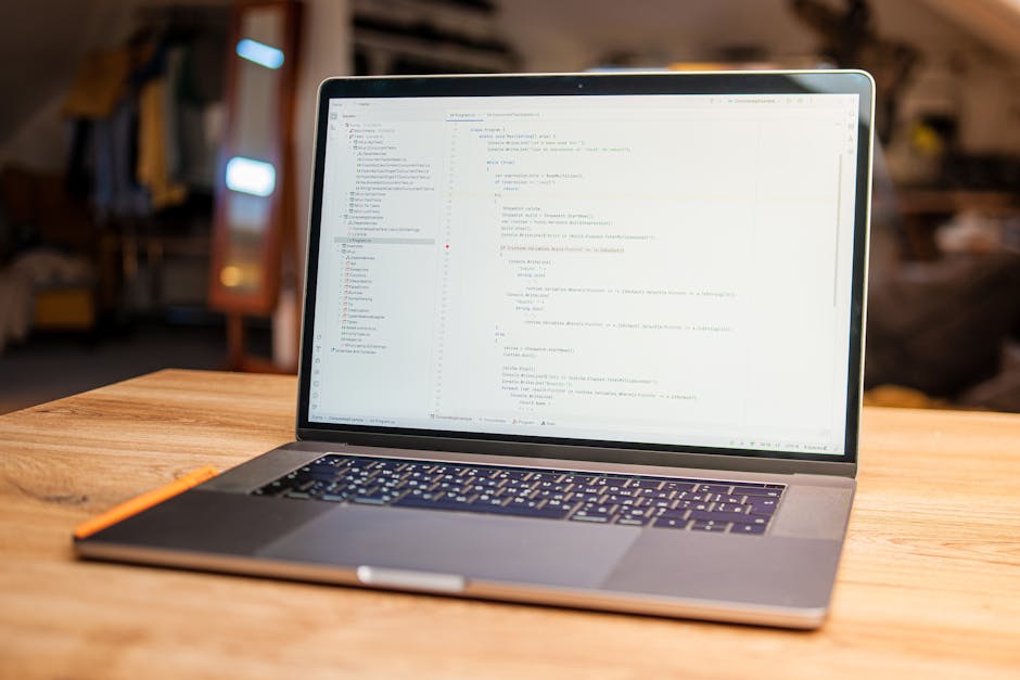 A silver laptop with a black keyboard sits open on a wooden desk in a well-lit indoor space, displaying lines of computer code on its screen. The desk appears smooth with a natural wood grain finish. 
