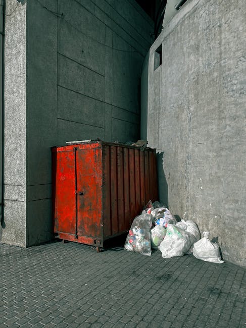 A pile of discarded rubbish on a gravel surface outdoors, consisting of black and white plastic bags filled with waste, an old, worn car tyre leaning against the heap, and a yellow plastic container. 