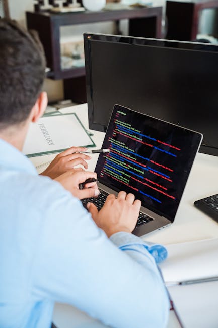 A person working at a desk is seen from behind, with short dark hair, wearing a light blue shirt. They are using a laptop with a black screen displaying lines of multicolored code in blue, red, yellow